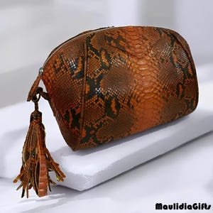 May include: A brown and orange snakeskin-patterned cosmetic bag with a zipper closure and a decorative tassel. The bag has a rounded shape and is made of a textured material. The tassel is brown and has a fringed design.