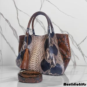 May include: A snakeskin-patterned handbag with a matching shoulder strap. The bag features a mix of brown, tan, and navy blue tones, with a tassel detail. The handles are brown, and the bag is set against a marble background.