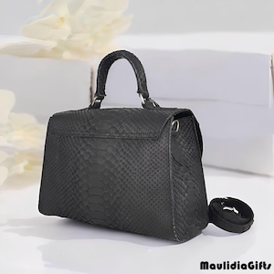 May include: A black handbag with a textured snakeskin pattern. The bag has a top handle, a shoulder strap, and a flap closure. The bag is sitting on a white surface.