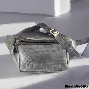 May include: A silver fanny pack with a textured, snakeskin-like pattern. The bag has a zippered main compartment and a smaller front pocket. A gold-colored buckle secures the adjustable strap. The bag is on a white surface.