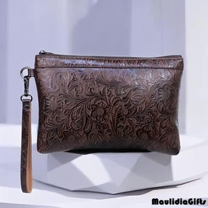 May include: A brown leather clutch with a floral embossed design. The clutch has a zipper closure and a wrist strap. The wrist strap is a lighter shade of brown. The clutch is rectangular and sits on a white surface.