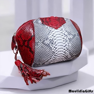 May include: A small, oval-shaped clutch with a red and silver snakeskin pattern. The clutch has a red zipper and a matching red tassel. The bag is sitting on a white surface. The brand name "MaulidiaGifts" is printed in black.