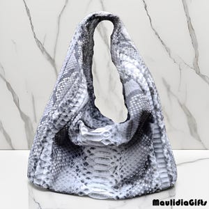 May include: A gray and white snakeskin-patterned handbag with a large, open handle. The bag is made of a soft, flexible material and has a rounded shape. The bag is sitting on a white marble surface.