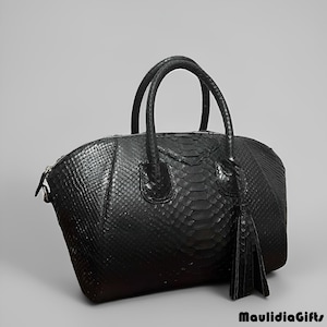 May include: A black leather handbag with a textured snakeskin pattern. The bag has two rounded handles, a zipper closure, and a decorative tassel. The bag is approximately 12 inches wide and 8 inches tall.