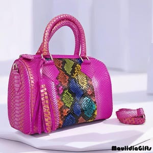 May include: A bright pink handbag with a snakeskin texture. The center panel features a colorful snakeskin pattern in shades of blue, green, yellow, and orange. Includes a matching strap and tassel.