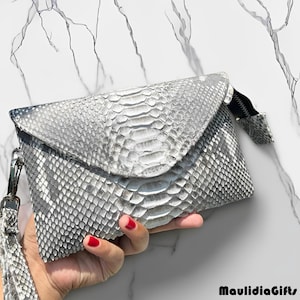 May include: A silver and white clutch with a snakeskin pattern. The clutch has a flap closure and a wrist strap with a tassel. The bag is made of a textured material and has a zipper closure. The overall design is sleek and stylish.