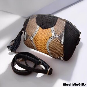 May include: A snakeskin-patterned clutch with a black leather tassel and a detachable black strap. The clutch features a zipper closure and a color palette of brown, tan, black, and orange. The brand name "MaulidiaGifts" is visible in the bottom right corner.