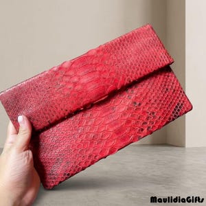 May include: A red clutch with a snakeskin pattern. The clutch is rectangular with a fold-over flap. The texture of the material is visible. The brand name "MaulidiaGifts" is in the bottom right corner.