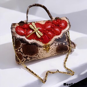 May include: A snakeskin-patterned handbag with a gold chain strap and handle. The flap is adorned with red beads and gemstones, and a gold dragonfly charm. The bag has a gold clasp.