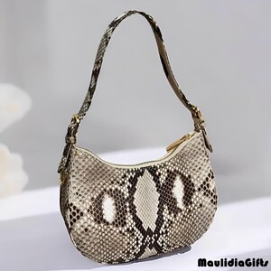 May include: A small, crescent-shaped handbag with a snakeskin pattern in shades of gray, brown, and white. The bag has a single handle, a gold-colored zipper, and gold-toned hardware. The bag is a stylish accessory.