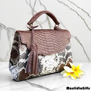 May include: A handcrafted handbag with a snakeskin pattern in shades of brown, gray, and white. The bag features a top handle, a flap closure, and a decorative tassel. A yellow and white flower rests beside the bag. The brand name "MaulidiaGifts" is at the bottom.