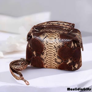 May include: A small, square handbag made of brown and tan snakeskin. The bag has a zipper closure and a detachable strap. The snakeskin pattern is visible on the exterior of the bag. The bag is sitting on a white surface.