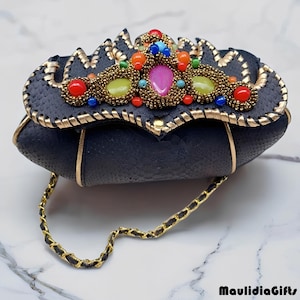 May include: Black clutch with a decorative top featuring colorful gemstones and gold accents. The clutch has a gold chain strap and is made of a textured material. The clutch is a unique accessory.