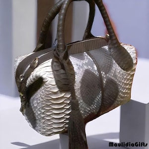 May include: A handbag with a snakeskin pattern in shades of white, gray, and brown. The bag has two handles and a zipper closure. The bag's design features a textured, scaled pattern, and a tassel. The brand name "MaulidiaGifts" is visible.