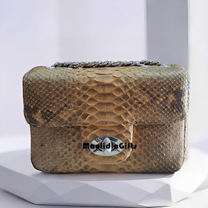 May include: A small, rectangular handbag with a snakeskin pattern in shades of brown and tan. It features a silver chain strap and a silver clasp with the text "maulidiaGifts". The bag is sitting on a white surface.