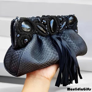 May include: Black clutch with a textured snakeskin pattern. The top of the clutch is embellished with black beads and large, dark stones. A fringe of black leather hangs from the bottom. The clutch is held in a hand.