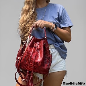 May include: A red leather bucket bag with a snakeskin pattern. The bag has a drawstring closure, a shoulder strap, and a top handle. The bag also has tassels and a white tag.