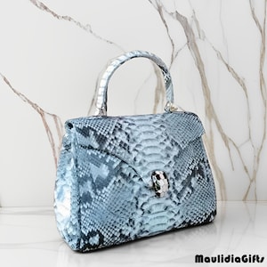 May include: A light blue handbag with a snakeskin pattern. The bag has a top handle and a flap closure with a decorative clasp. The bag is sitting on a white surface with a marble background. The text "MaulidiaGifts" is in the bottom right corner.