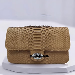 May include: A tan rectangular handbag featuring a textured snakeskin pattern across the exterior. The bag includes a silver-toned metal turn-lock closure on the front flap and a metallic chain strap. The text MaulidiaGifts is printed in black.