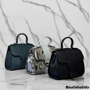 May include: Three leather handbags in shades of green, gray, and black. Each bag has a top handle and a flap closure. The bags are displayed on a white marble surface.