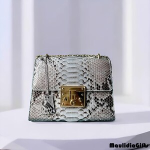 May include: A square-shaped handbag with a snakeskin pattern in shades of gray and white. The bag features a gold-colored clasp and chain strap. A small tag hangs from the front. The bag is sitting on a white surface.