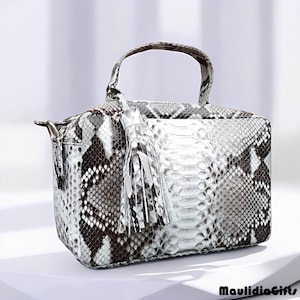 May include: A rectangular handbag with a snakeskin pattern in shades of white, gray, and black. It features a top handle, a zipper closure, and a decorative tassel. The bag is sitting on a white surface.