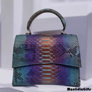 May include: A small, colorful handbag with a top handle. The bag features a snakeskin pattern in shades of teal, purple, and orange. The handle is a darker teal color. The bag has a rectangular shape with a flap closure. The text "MaulidiaGifts" is in the bottom right corner.