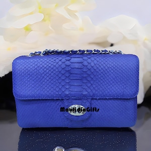 May include: A cobalt blue handbag with a chain strap. The bag is made of a textured material, possibly snakeskin, and features a silver clasp. The text "MaulidjaGifts" is visible on the front of the bag.