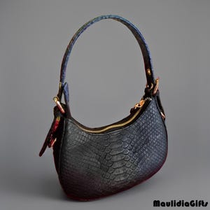 May include: Black handbag with a textured, snake-like pattern. The bag has a curved shape, a gold zipper, and a handle with a colorful floral and blue design. The bag is set against a gray background.
