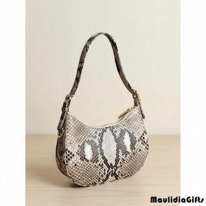 May include: A small, crescent-shaped handbag with a snakeskin pattern in shades of gray, black, and white. The bag has a single, adjustable strap and a gold-toned zipper closure. The bag is made of a textured material and is set against a neutral background.