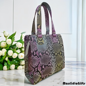 May include: A colorful tote bag with a snakeskin pattern in shades of purple, black, white, and yellow. The bag has two handles and a zipper closure. The bag is sitting on a marble surface.