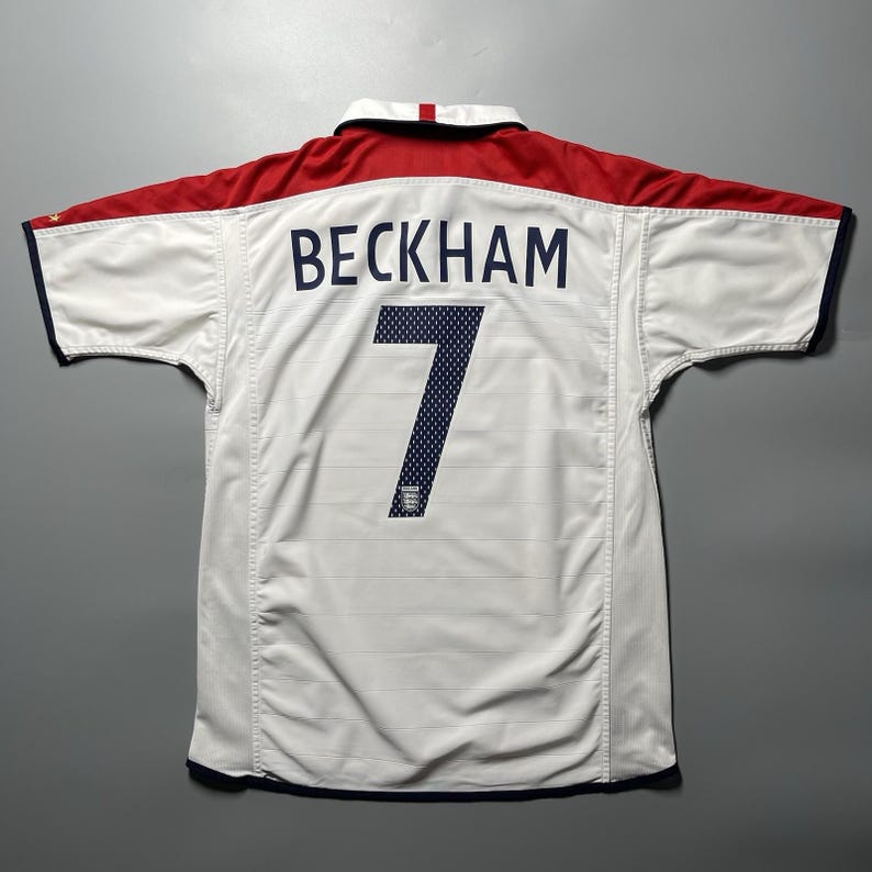 May include: White soccer jersey with red accents on the shoulders and collar. The name "BECKHAM" is printed across the back in navy blue, with the number "7" below. The jersey has a navy blue trim.