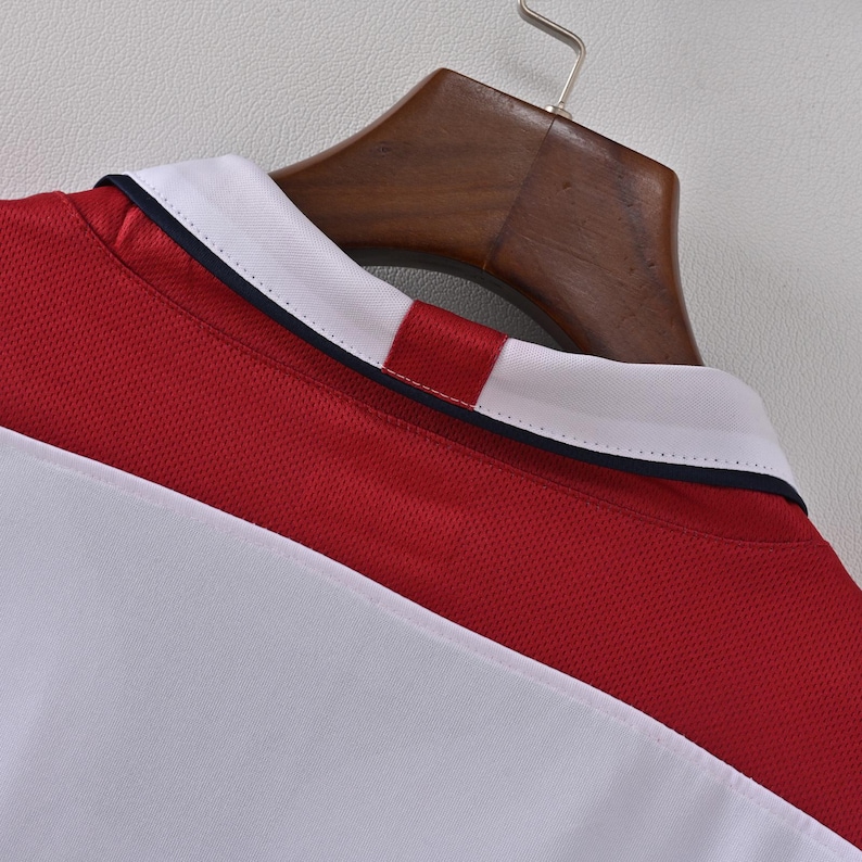 May include: A red and white athletic jersey with a white collar and a navy blue trim. The jersey is hanging on a wooden hanger. The fabric has a mesh texture.
