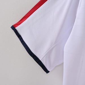May include: Close-up of a white athletic shirt sleeve with red and navy blue accents. The sleeve has a mesh panel for breathability and a navy blue trim. The shirt appears to be made of a lightweight, breathable fabric.