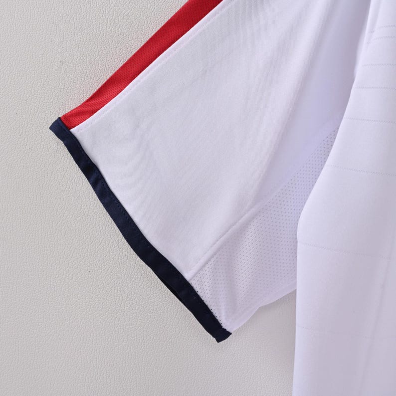 May include: Close-up of a white athletic shirt sleeve with red and navy blue accents. The sleeve has a mesh panel for breathability and a navy blue trim. The shirt appears to be made of a lightweight, breathable fabric.