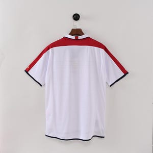 May include: White short-sleeved shirt with a red yoke and shoulder stripes. The shirt has a collared neckline and navy blue trim on the sleeves and hem. The shirt is hanging on a wooden hanger.