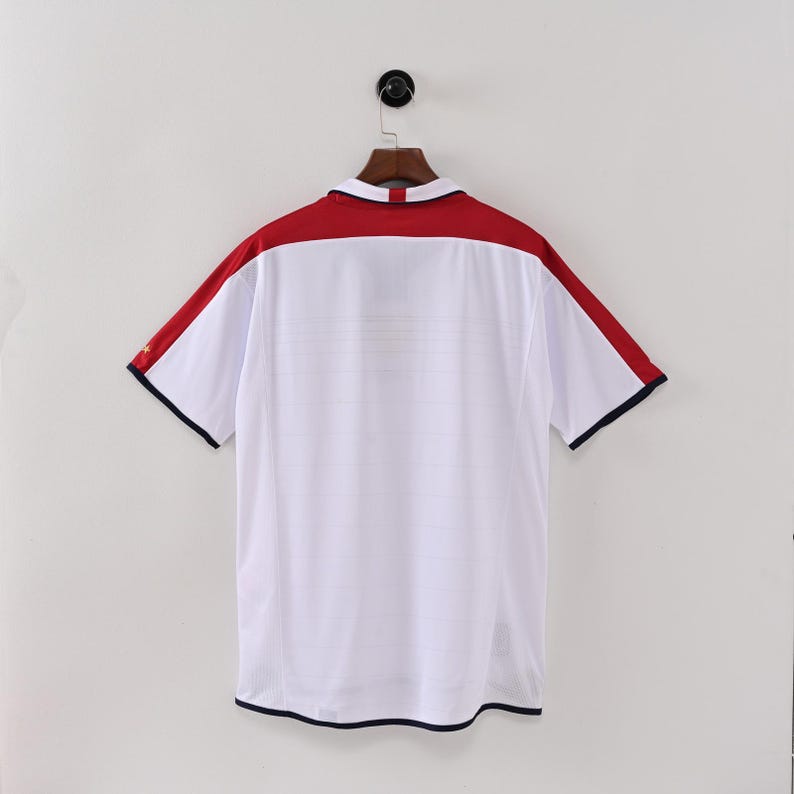 May include: White short-sleeved shirt with a red yoke and shoulder stripes. The shirt has a collared neckline and navy blue trim on the sleeves and hem. The shirt is hanging on a wooden hanger.