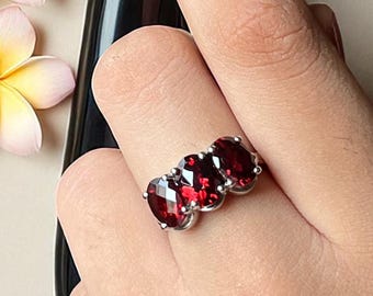 May include: A silver ring featuring three oval-cut deep red garnet gemstones arranged horizontally in a prong setting. The ring is worn on a finger, positioned next to a black-handled fork and three light pink and yellow plumeria flowers.