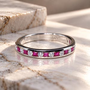 Könnte beinhalten: Ein silberfarbener Bandring mit einem abwechselnden Muster aus runden, klaren Steinen und quadratisch geschliffenen, leuchtend pinken Rubinen in Kanalfassung. Der Metallring hat eine polierte Oberfläche und eine gleichmäßige Breite.