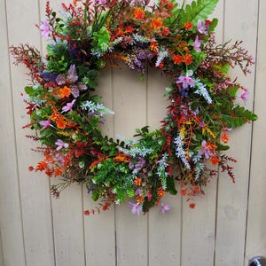 Puede incluir: Una vibrante corona floral con una variedad de flores y vegetación coloridas. La corona presenta flores naranjas, rosas, rojas y blancas, con hojas verdes en forma de helecho. La corona se exhibe en una puerta de madera de color claro.
