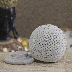 Op de afbeelding: Een witte stenen kaarsenhouder met een bolvormige deksel met een rasterpatroon en een ronde voet. Het object bevat een klein, rond, zilverkleurig theelichtje dat centraal op de voet is geplaatst en klaar is voor gebruik in huis.
