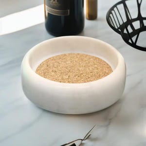 May include: A round white marble bowl featuring a smooth, polished exterior and a thick rim. The interior base of the bowl is lined with a circular, light brown cork insert, providing a textured contrast to the solid stone construction.