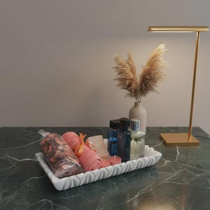 Puede incluir: Una bandeja rectangular blanca con borde festoneado contiene una bolsa transparente de popurrí seco, una toalla enrollada color coral con una cinta naranja, un jabón rectangular, dos velas redondas pequeñas y dos frascos de vidrio con líquido.