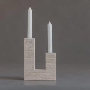 Puede incluir: Un portavelas moderno en forma de U, hecho de piedra beige claro. Sostiene dos velas blancas cónicas. El diseño es minimalista, sobre un fondo gris liso.