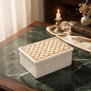 May include: A rectangular white marble box with a gold lattice design on the lid. The box sits on a green marble surface, suggesting a luxury item. The box is likely used for storing jewelry or small items.