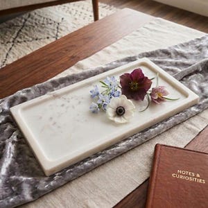 May include: A rectangular white marble tray with subtle grey veining. Resting on the tray are several fresh flowers, including a large white anemone with a dark centre, a deep purple hellebore, small blue star-shaped flowers, and a small patterned bud.