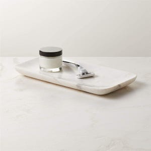 May include: A rectangular white marble tray with grey veining holding a small glass jar with a black lid containing white cream and a silver and white manual razor. The tray has smooth, rounded edges and a polished finish for a clean look.