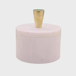 Puede incluir: Un recipiente cilíndrico hecho de mármol rosa pálido con un acabado liso y pulido. El recipiente cuenta con una tapa circular a juego rematada con un único mango metálico de color dorado, cónico y centrado en la parte superior de la tapa.