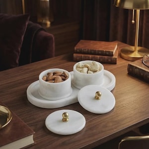 May include: White marble serving set with two lidded bowls filled with almonds and cashews, resting on a matching oval tray. The lids have gold-colored knobs. The set is on a dark wood surface.