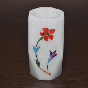 Puede incluir: Un portalápices cilíndrico de mármol blanco con un diseño floral incrustado en colores. La incrustación muestra una flor grande roja con centro azul, una flor más pequeña azul y roja, y hojas verdes, unidas por tallos negros finos.